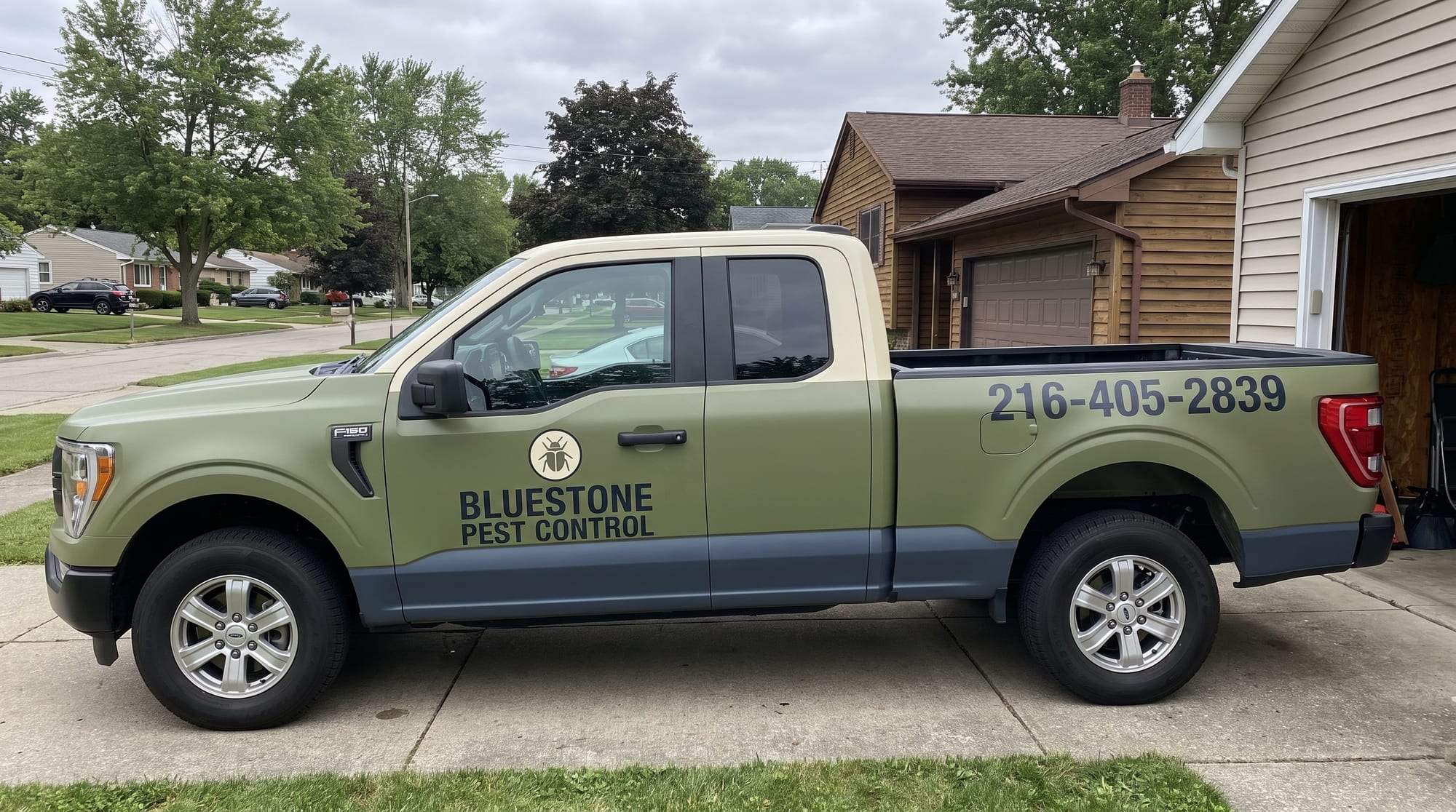 Bluestone Pest Control pickup truck, olive green with a cream roof and dark slate lower panels, the beetle logo and company name on the door and 216-405-2839 on the bed, parked on a concrete driveway beside a house with an open garage